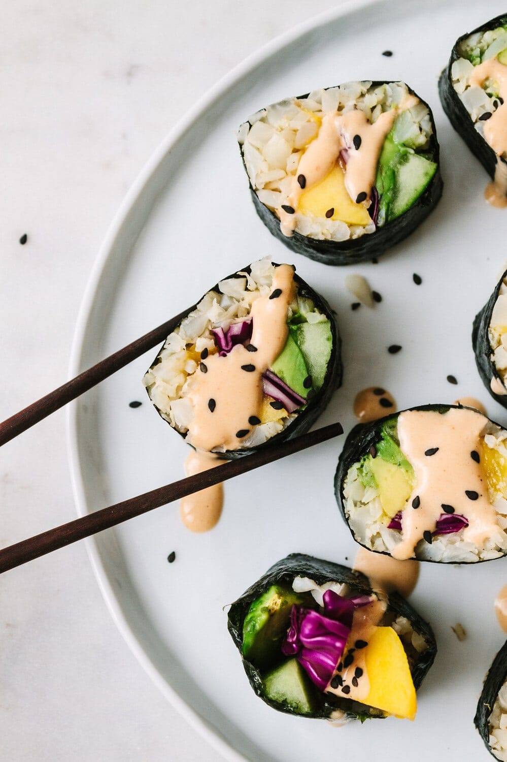 top down view of chopsticks holding mango avocado cauliflower rice sushi rolls drizzled with spicy dynamite sauce on a plate.