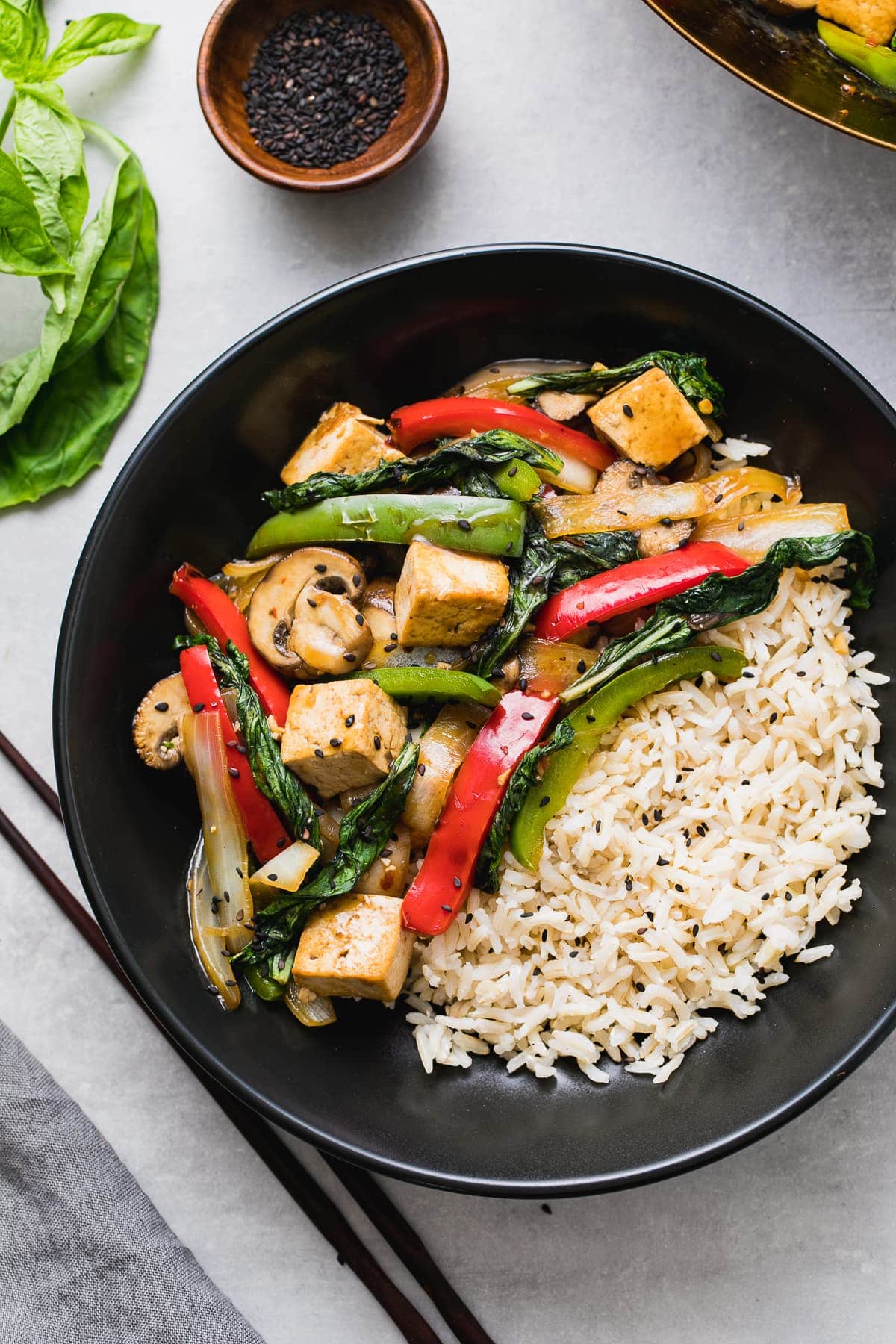 top down view of a serving of thai basil stir fry with tofu and veggies, and rice on the side, in a black bowl.