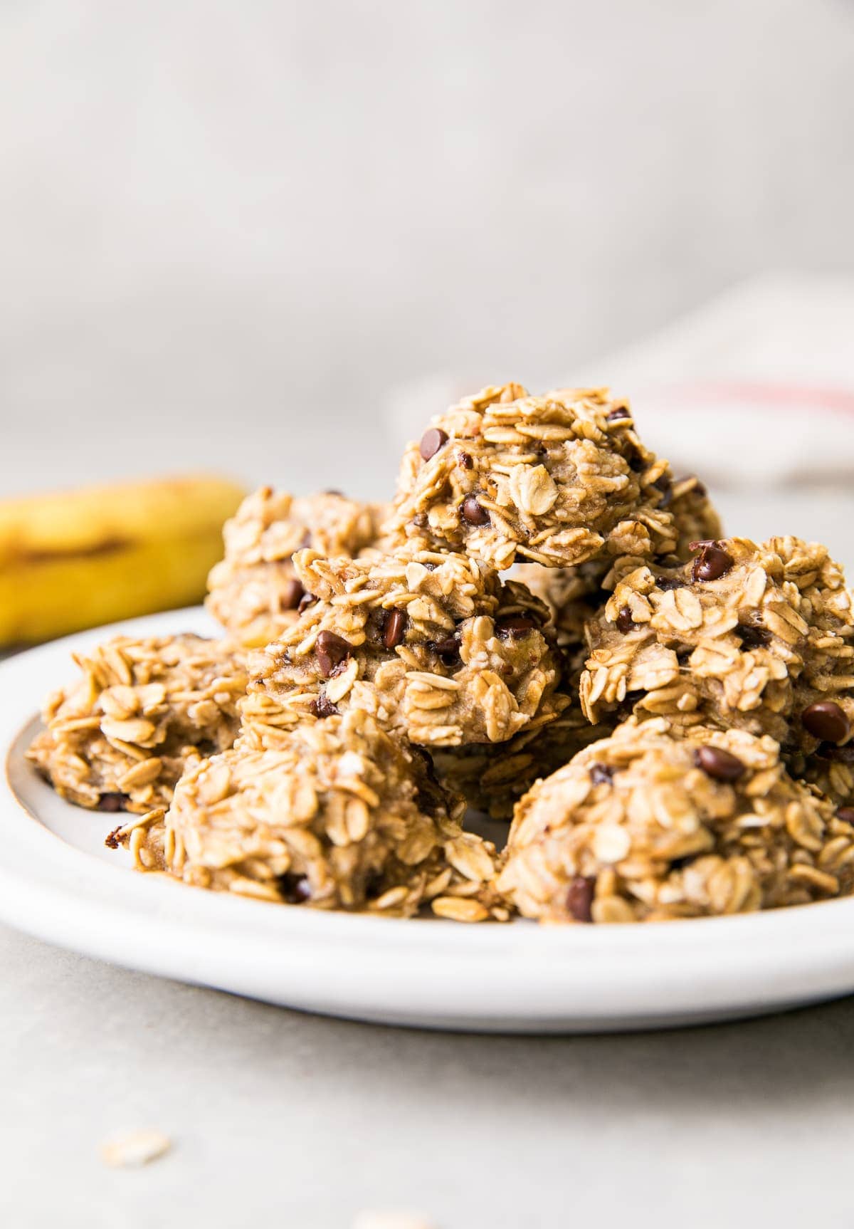 side angle view of group of stacked banana oatmeal chocolate chip cookies on a white plate with items surrounding.