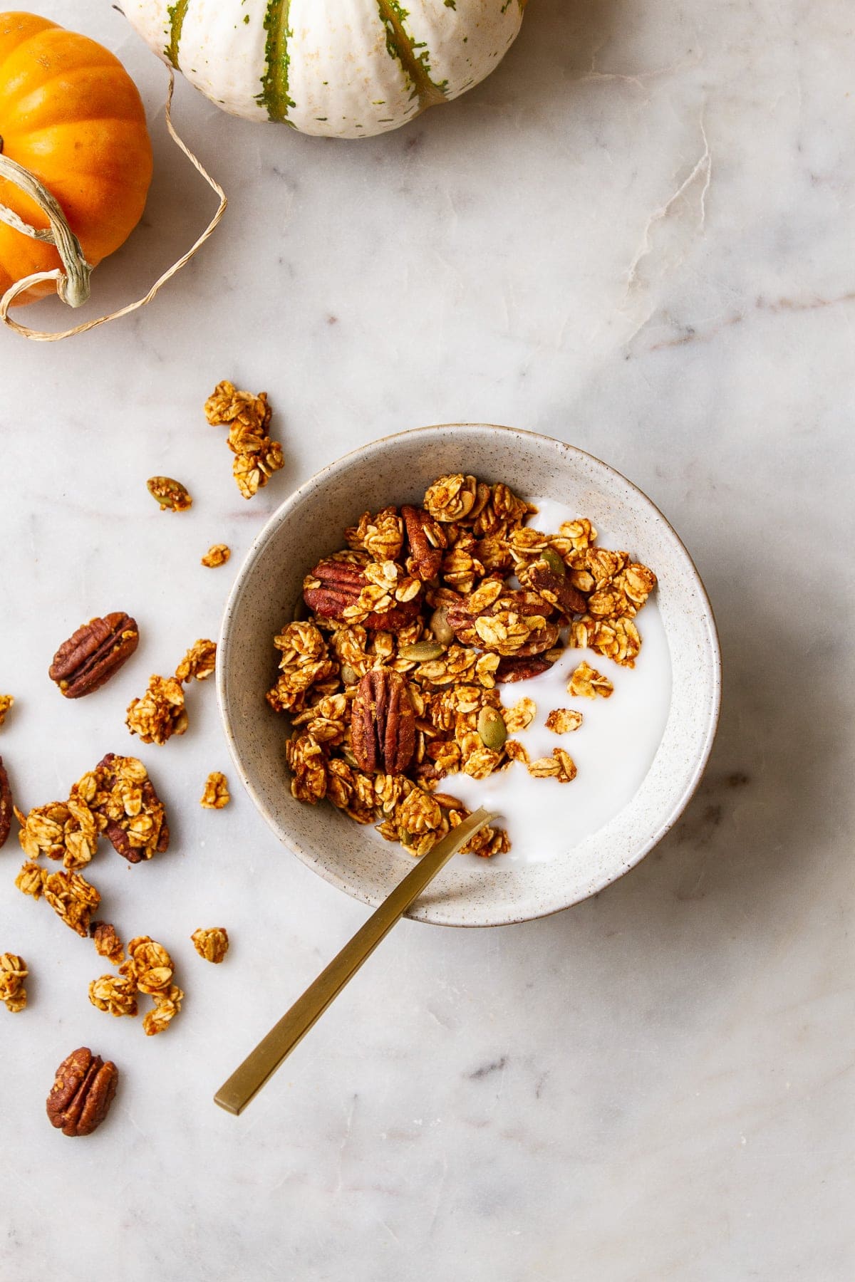 top down view of a bowl with a serving of coconut yogurt and pumpkin granola with a gold spoon surrounded by items.