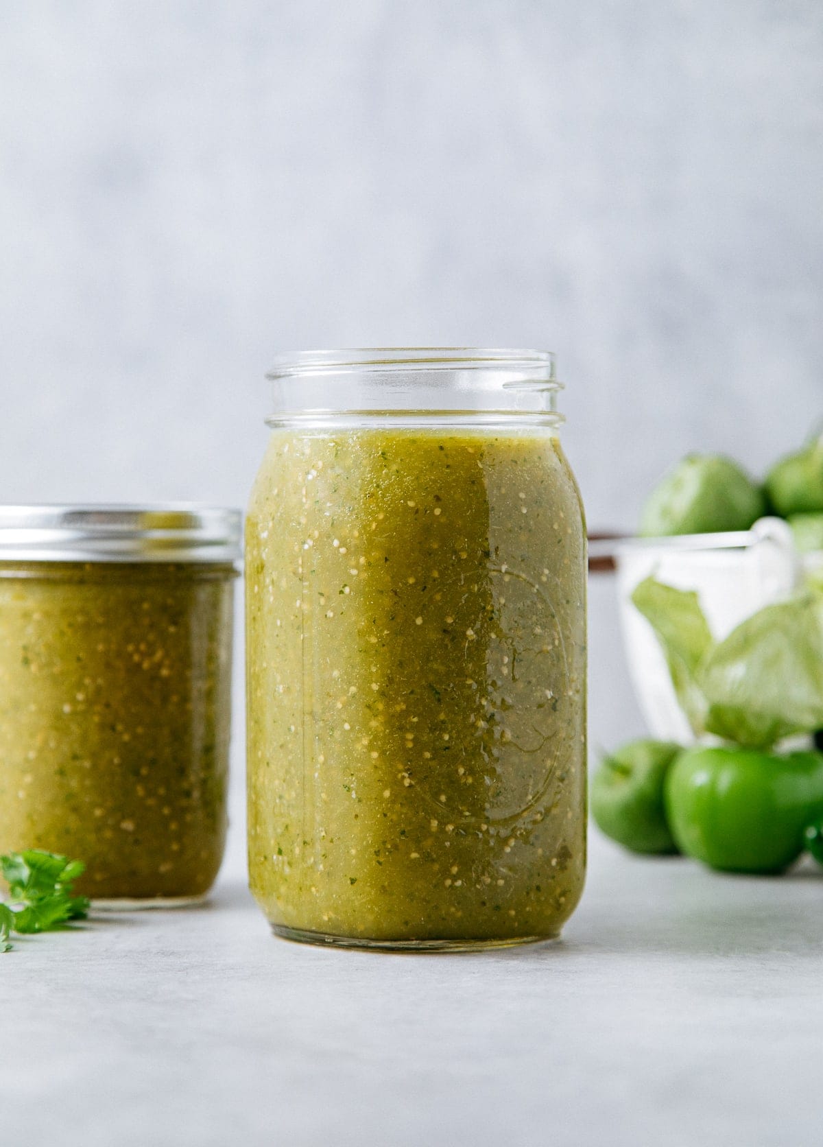 head on view of glass jar filled with homemade tomatillo salsa verde and items in background.