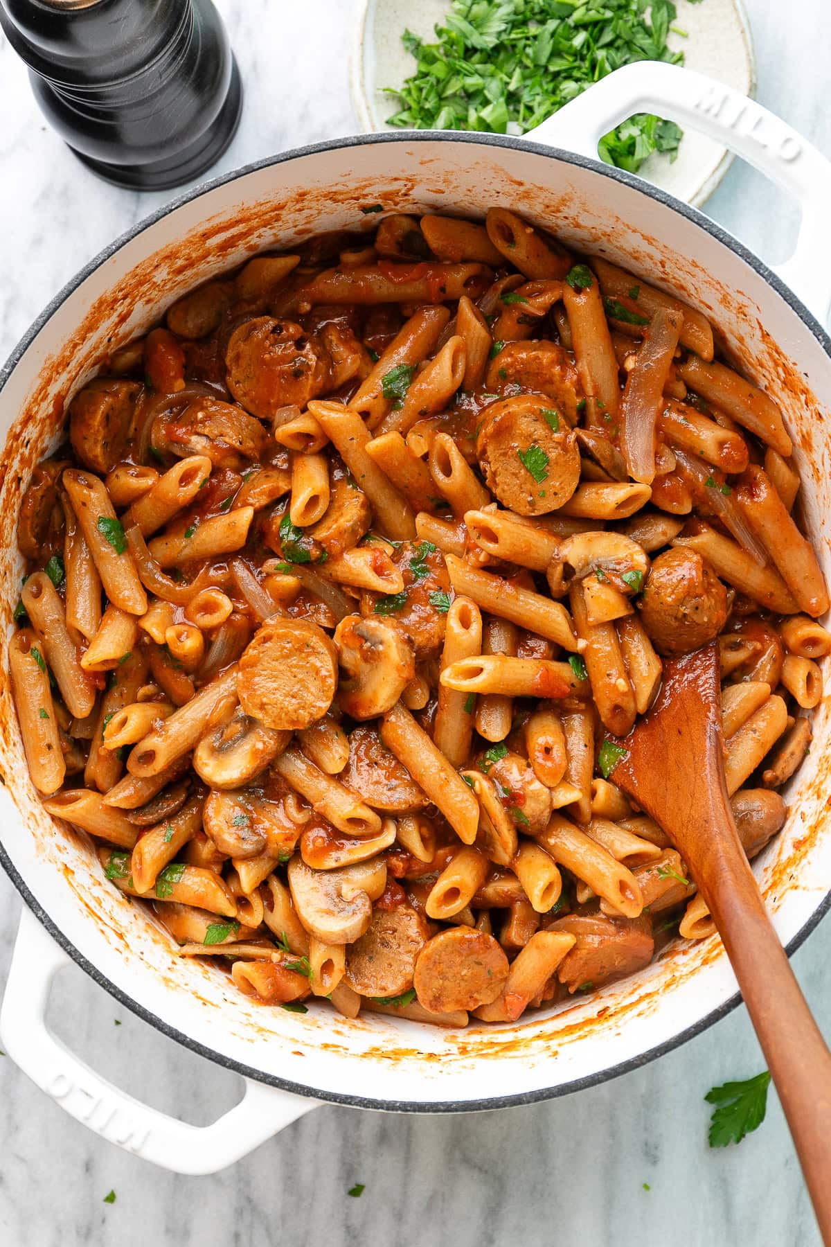 top down view of freshly made vegan sausage pasta in a white pot with wooden spoon.