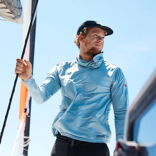 A man in a long sleeved blue shirt and a black baseball cap reading Florence Marine X stands on a boat holding onto the rigging.