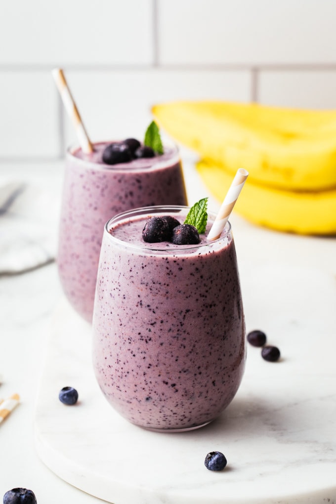 head on view of glass full of blueberry banana smoothie with straw and items surrounding.