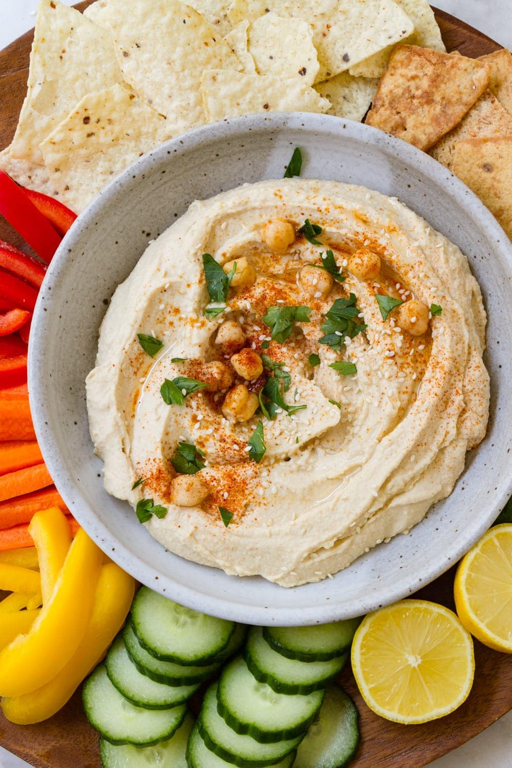 top down view of best authentic hummus in a bowl topped with parsley, chickpeas and paprika, and surrounded by chips and sliced vegetables for dipping.