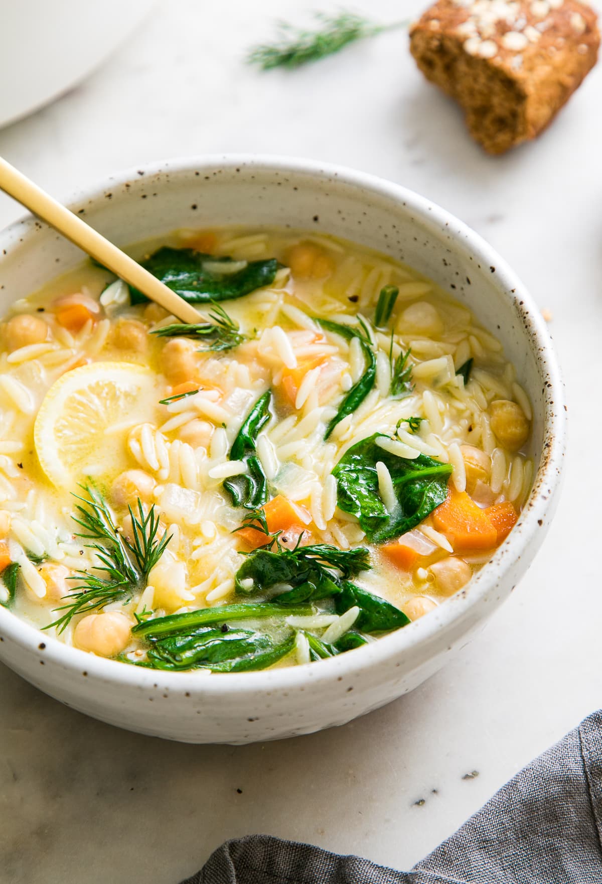 side angle view of bowl full of lemon chickpea orzo soup with gold spoon and items surrounding.