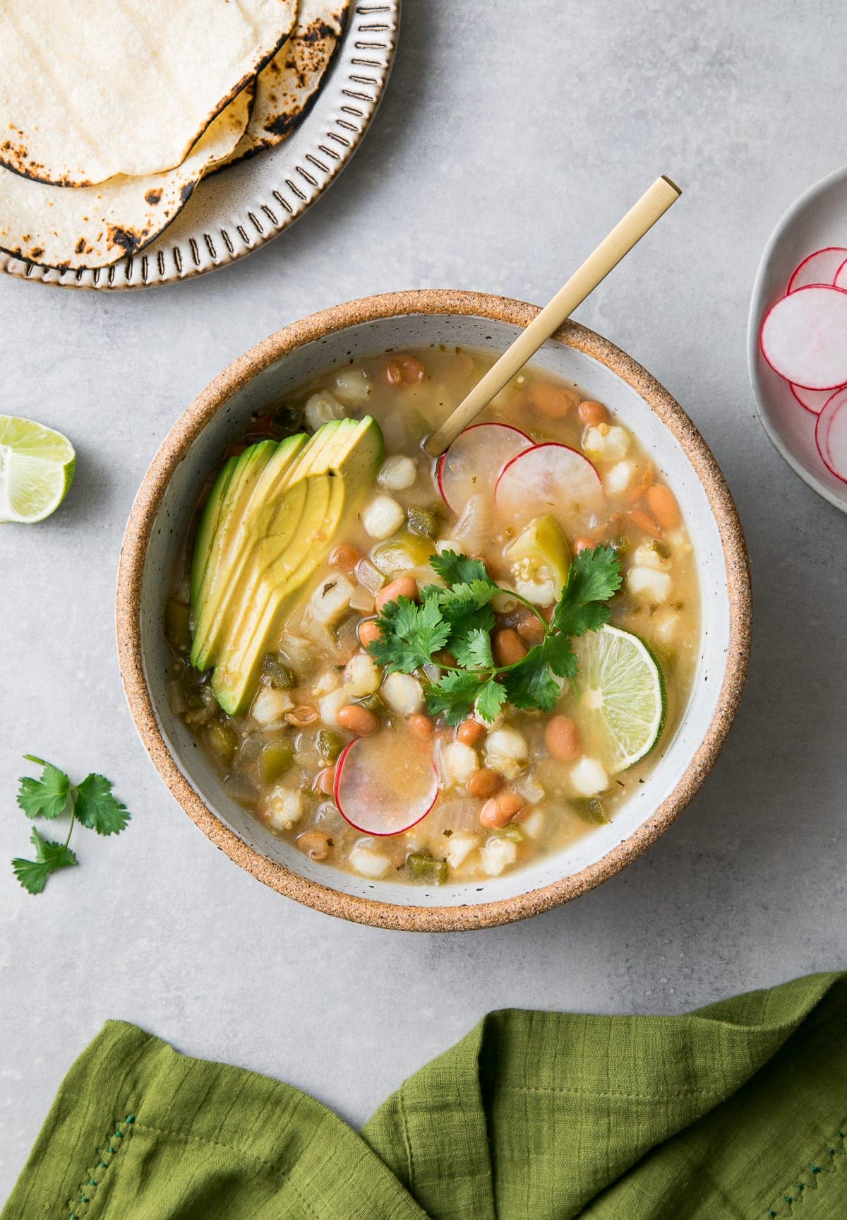 top down view of bowl with posole verde with spoon and items surrounding.