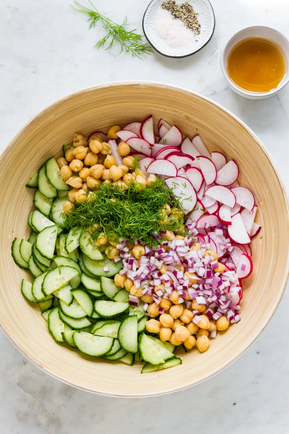 top down view of the ingredients for radish cucumber salad in a mixing bowl with the dressing in a small bowl to the upper ride side