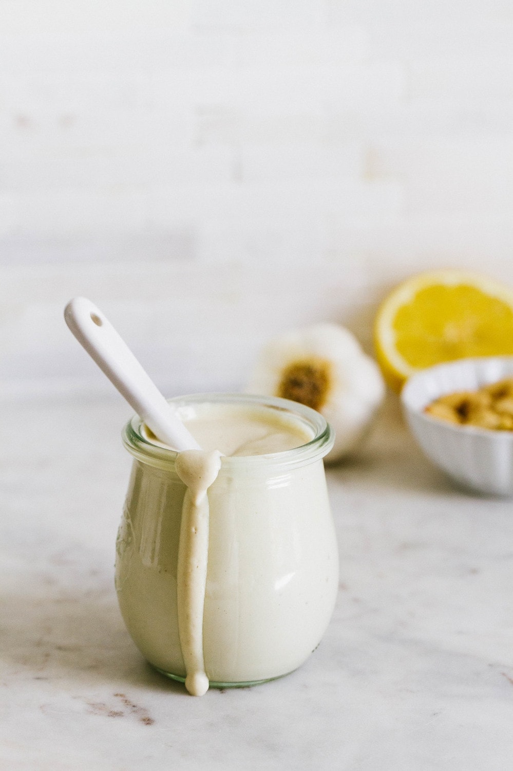 straight on view of vegan aioli in a glass jar with spoon sticking out, in the background is a small bowl of cashews, garlic bulb and half of lemon