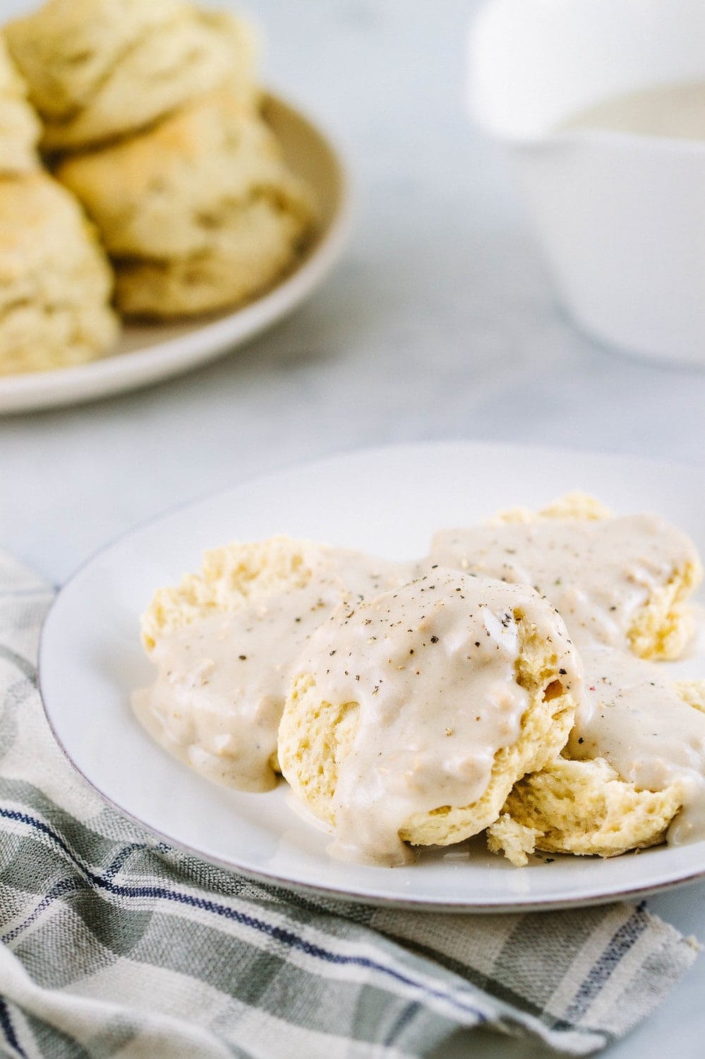 head on view of vegan biscuits topped with gravy on a small white plate with a plate of stacked biscuits and gravy bowl in the background.